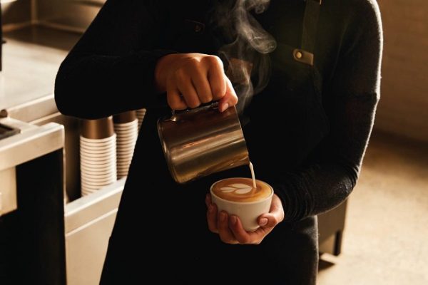 barista-wearing-black-pouring-cup-of-coffee-metal-milk-jug